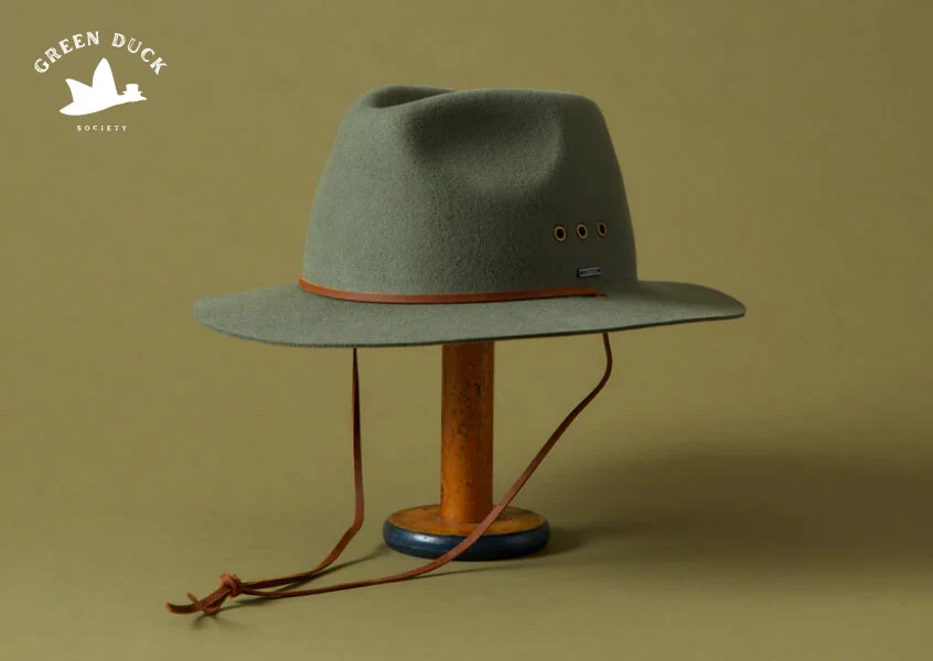 Minimalist product showcase of a handcrafted green felt hat with leather band and chin strap, styled on a wooden stand against a muted background, representing clean and modern e-commerce web design aesthetics.