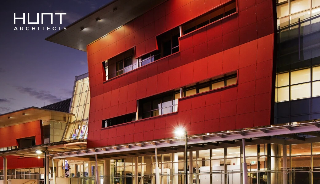 Modern architectural building with bold red exterior and glass elements at dusk, featuring Hunt Architects branding, showcasing contemporary design aesthetics in an architecture firm website